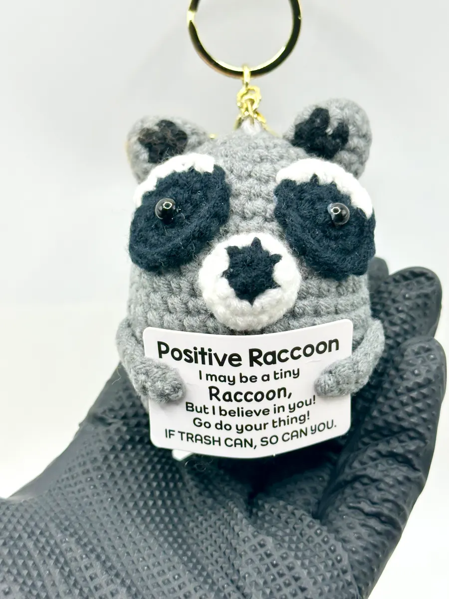 Grey crochet raccoon keychain holding a Positive Raccoon card